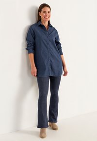 Takkipaita, jossa on kauluksen pääntie, pitkät hihat ja napitus, yhdistetty tumma denimpohjaisiin ryhdikkäisiin housuihin ja vaaleanbeigeihin nilkkabootseihin.