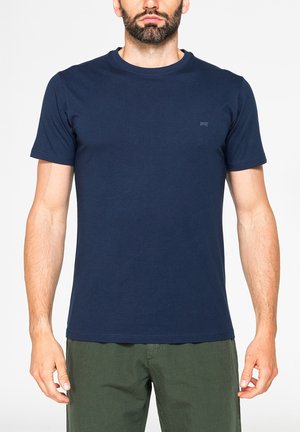 Uomo con barba che indossa una maglietta blu navy tinta unita a maniche corte e pantaloni verde oliva, in piedi davanti a uno sfondo bianco.