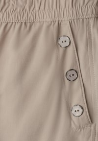 Beige fabric with elastic waistband and three vertically aligned round buttons with two holes each on angled flap.
