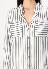 Chemise blanche à manches longues avec des rayures noires verticales, des rayures horizontales contrastantes sur deux poches poitrine et des boutons beiges sur le devant.