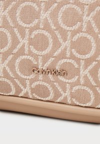  Bézs Calvin Klein kézitáska, texturált anyagból készült, ismétlődő logómintával és sima alappal. Arany színű kiegészítők láthatók.