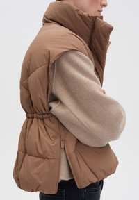 Braune, gepolsterte Weste mit hohem Kragen, Zugband in der Taille und seitlichem Reißverschluss. Tragen über einem hellbeigen Pullover mit strukturierter Oberfläche.