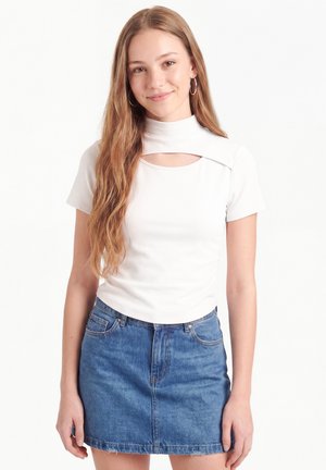 Jeune femme aux longs cheveux portant un haut blanc à manches courtes avec une découpe et une jupe en jean bleue, debout sur un fond blanc.