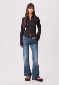 Chemise noire ajustée à manches longues, cintrée à la taille. Jean en denim bleu évasé avec un délavage et des coutures claires. Sandales à plateforme marron.