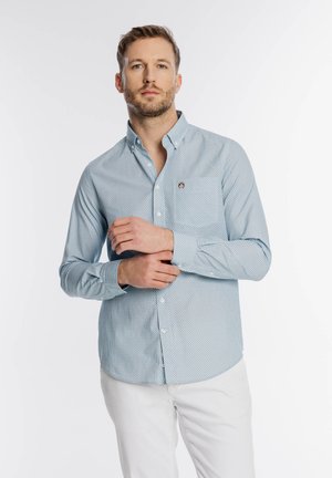 Mężczyzna poprawiający mankiet jasnoniebieskiej, wzorzystej koszuli zapinanej na guziki, ubrany w białe spodnie, stojący na tle jednolitej białej ściany.