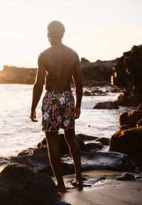 Heren zwemshorts met een donker bloemenpatroon met roze en witte bloemen. De stof lijkt lichtgewicht te zijn. Zicht van achteren nabij het water.
