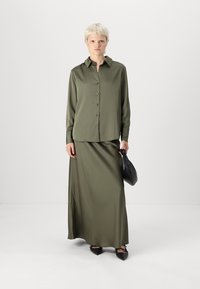 Chemise olive verte à manches longues avec des boutons ; jupe assortie jusqu'aux chevilles. Tissu doux et lisse ; sac noir plat ; chaussures noires à bout pointu.