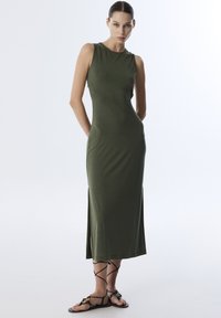 Olijfgroen mouwloos maxi-jurk met een aansluitende pasvorm en zijsplitten. Glad stof met een lichte glans. Platte, gespsandalen met decoratieve accenten.