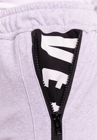 Ljust grå träningsoverallsbyxor med en svart dragkedja som avslöjar en svart och vit logotyp inuti. Mjuk tyg med resår i midjan.