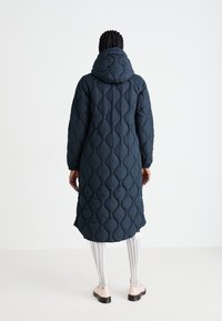 Donkerblauwe pufferjas met een gewatteerd patroon, capuchon en lange mouwen. Bereikt de middenkuit met zijsplitten, gedragen met een patroonlegging en schoenen.