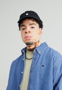 Camisa de mezclilla con cuello abotonado, color azul claro, logo en el pecho. Gorra negra, camiseta verde debajo, collar de cadena de plata visible.