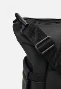 Sac à épaule noir en matériau lisse et imperméable. Doté d'une sangle texturée et d'un crochet en métal. Design simple avec quincaillerie durable.