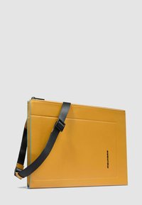 Borsa a tracolla rettangolare in pelle giallo senape con tracolla nera regolabile e bordatura blu, esposta su sfondo grigio chiaro uniforme.