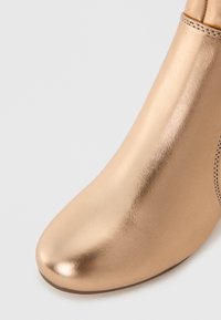Botín metálico dorado con textura lisa, punta redondeada y mínimos detalles de costura. Diseño elegante con una silueta ajustada.