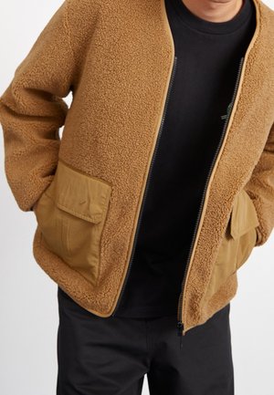 Homme portant une veste polaire beige avec de grandes poches avant sur une chemise noire et un pantalon noir, les mains dans les poches de la veste.