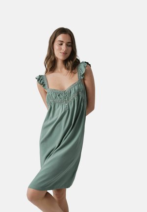 Jeune femme aux cheveux bruns mi-longs portant une robe verte sans manches avec des bretelles à volants, debout devant un fond uni.