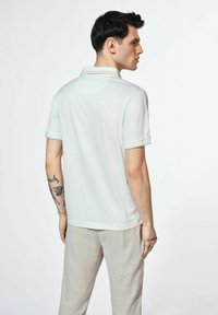 Biała koszulka polo z krótkimi rękawami, z klasycznym kołnierzykiem. Materiał wydaje się gładki i lekki. Noszona z jasnymi spodniami.