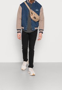 Giacca bomber a blocchi di colore denimm e beige, t-shirt grigia, jeans neri e sneakers chiare. Borsa a tracolla beige con cerniera.