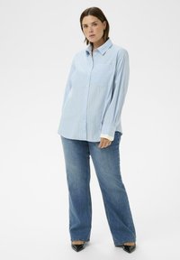 Niebieska koszula w paski z długim rękawem, z pojedynczą kieszenią na piersi; zestawiona z szerokimi niebieskimi dżinsami i czarnymi butami z zaostrzonym noskiem.