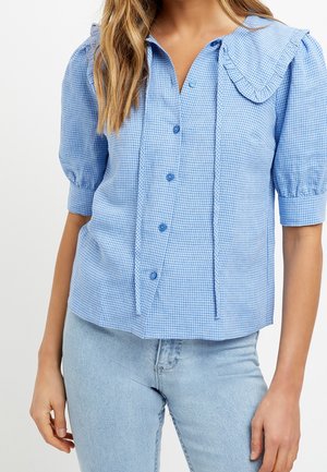 Femme portant un chemisier à manches bouffantes en vichy bleu clair avec un col à volants, associé à un jean en denim délavé clair.