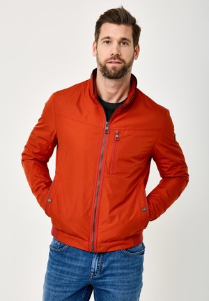 Mann mit Bart, der eine rote Jacke mit Reißverschluss und blaue Jeans trägt, steht mit den Händen in den Taschen vor einem einfarbigen weißen Hintergrund.