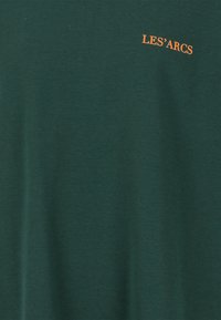 Tumman vihreä puuvillapaita, jossa on hillitty oranssi "LES'ARCS" -logo brodeerattuna vasemmassa yläkulmassa. Pehmeä rakenne, rento muotoilu.