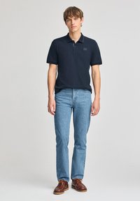 Polo de color azul marino con mangas cortas, un cuello y un parche texturizado; combinado con jeans de color azul claro y zapatos marrones con cordones.
