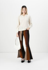 Vrouw in een crèmekleurige gebreide trui, multicolore bruine patchwork flared broek, zwarte schoenen, met een witte handtas, staande op een witte achtergrond.