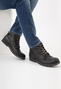 Black leather ankle boots with gray laces, flat rubber sole, and stitching detail. Styled with blue jeans on a white background.