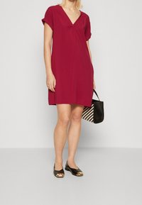 Robe courte à manches courtes couleur bordeaux avec un col en V et un détail plissé, associée à des ballerines noires ornées d'accents dorés et un sac à main à motifs.