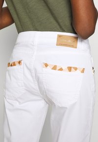 Vita bomullbyxor med mönstrade bakfickor som har geometriska former i persika och brunt. Läderetikett på midjan med varumärkesnamn.