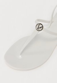 Valge plastikust flip-flop, kitsaste rihmade ja ümmarguse logo aktsendiga. Sile tekstuur ja minimalistlik disain, millel on lame tald.