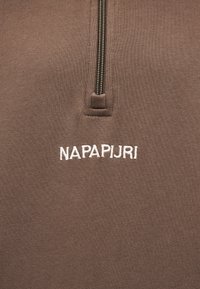 Καφέ ύφασμα με κάθετο φερμουάρ πάνω από λευκό κεντητό κείμενο «NAPAPIJRI» στην περιοχή του στήθους ενός ενδύματος.