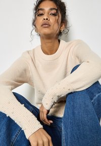 Hellrosa gestrickter Pullover mit Perlenakzenten an den Ärmeln, gerippten Bündchen und Rundhalsausschnitt, kombiniert mit blauen Jeans, sitzend vor einem neutralen Hintergrund.