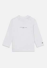 Weißes Langarmbabyschirt mit dem "Tommy Hilfiger EST. 1985" Logo auf der Brust und kleinem Flaggenlogo am Bündchen des linken Ärmels.