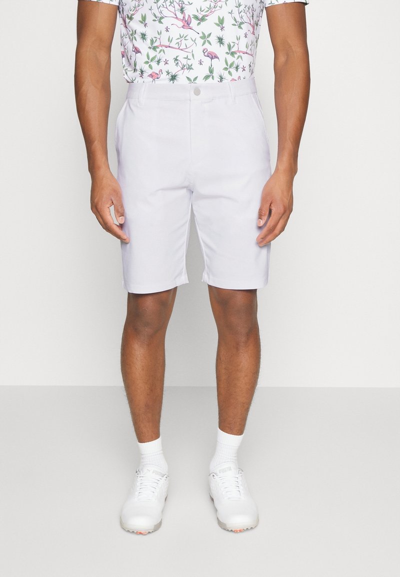 Homme portant un short blanc sur mesure, des chaussures et chaussettes de sport blanches, et une chemise à manches courtes avec un motif de flamant rose et de feuilles vertes.
