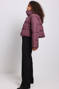 Trumpas pūkinis švarkas giliai violetinės spalvos, su aukštai kilpiniu kaklu, didelėmis horizontaliomis siūlėmis ir priekine užtrauktuku. Derinamas su juodomis kelnėmis.