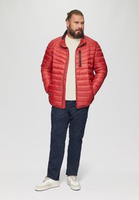 Rode gewatteerde jas met zwarte accenten, ritsluiting en zijzakken, gedragen over een lichte trui, gecombineerd met marineblauwe cargobroek en sneakers.