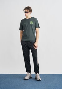 T-shirt de manga curta verde escura com estampado verde néon, combinada com calças de treino pretas com riscas laterais cinza claro e ténis cinzentos.