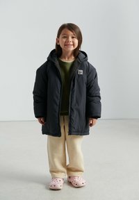 Tumehall kapuutsiga pufferjakk, kantud üle rohelise kampsuni ja helebeežide dressipükste. Kaasas roosad augulised klotsid.