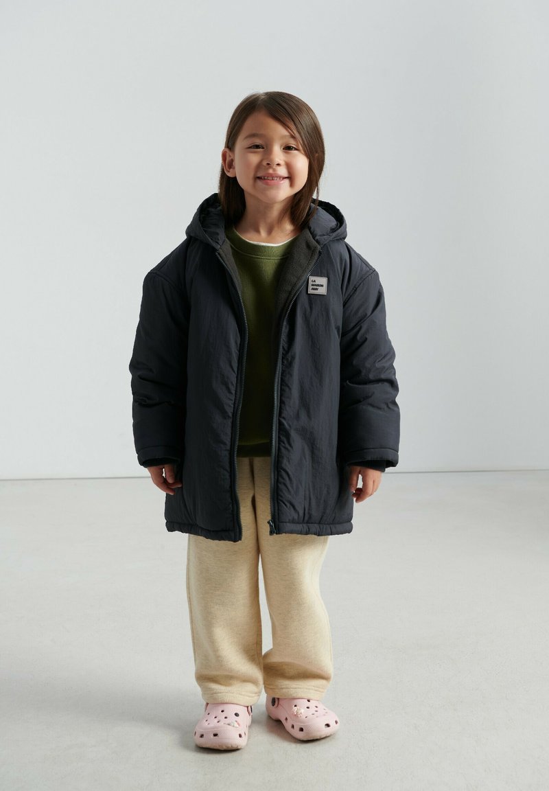 Tumehall kapuutsiga pufferjakk, kantud üle rohelise kampsuni ja helebeežide dressipükste. Kaasas roosad augulised klotsid.