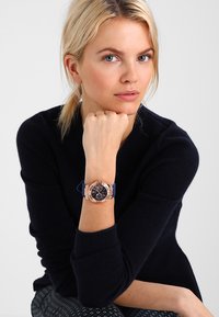 Lyxig analog klocka med armband i marinblå silikon och boett i ros�éguldton, med en svart urtavla och kronografdetaljer.
