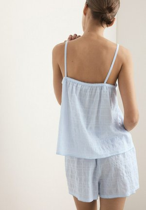 Femme aux cheveux attachés portant un haut sans manches bleu clair et un short assorti, vue de dos, debout contre un mur blanc.