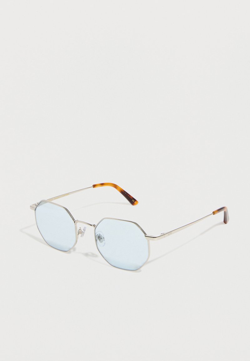 Οκταγωνικά γυαλιά ηλίου με ασημένιο πλαίσιο, φακούς σε ανοιχτό μπλε και καφέ άκρα να αγκαλιάζουν τα αυτιά, σε λευκό φόντο.