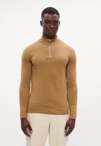 Maglione lavorato a maglia marrone con colletto alto, chiusura con zip e polsini a costine. Texture liscia, design aderente e dettagli di branding minimi.