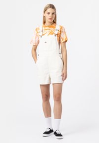 Hvite denim-dresser med justerbare stropper, kombinert med en oransje tie-dye skjorte, satt mot en ensfarget bakgrunn, med svarte sko og hvite sokker.