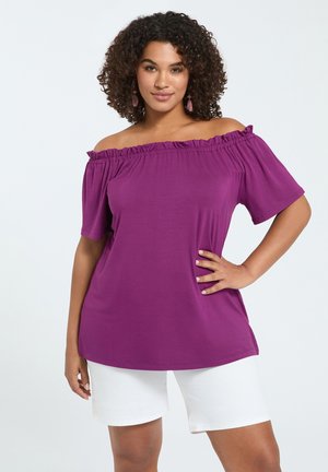 Frau mit lockigem Haar, die ein lila schulterfreies Oberteil und weiße Shorts trägt und mit einer Hand in der Hüfte vor weißem Hintergrund posiert.