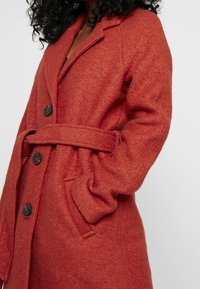 Cappotto in lana testurizzata di colore ruggine, con vita accentuata da una cintura, colletto a revers e grandi bottoni neri. Presenta tasche laterali e una silhouette morbida.