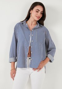 Camicia a bottoni con righe verticali blu e bianche, taschino sul petto, maniche arrotolate e una vestibilità morbida. Realizzata in un tessuto morbido.