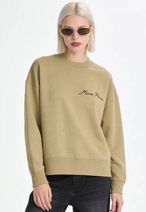 Giovane donna con capelli biondi che indossa una felpa marrone chiaro stampata con "Mira Paris" e jeans scuri, con occhiali da sole con montatura dorata.